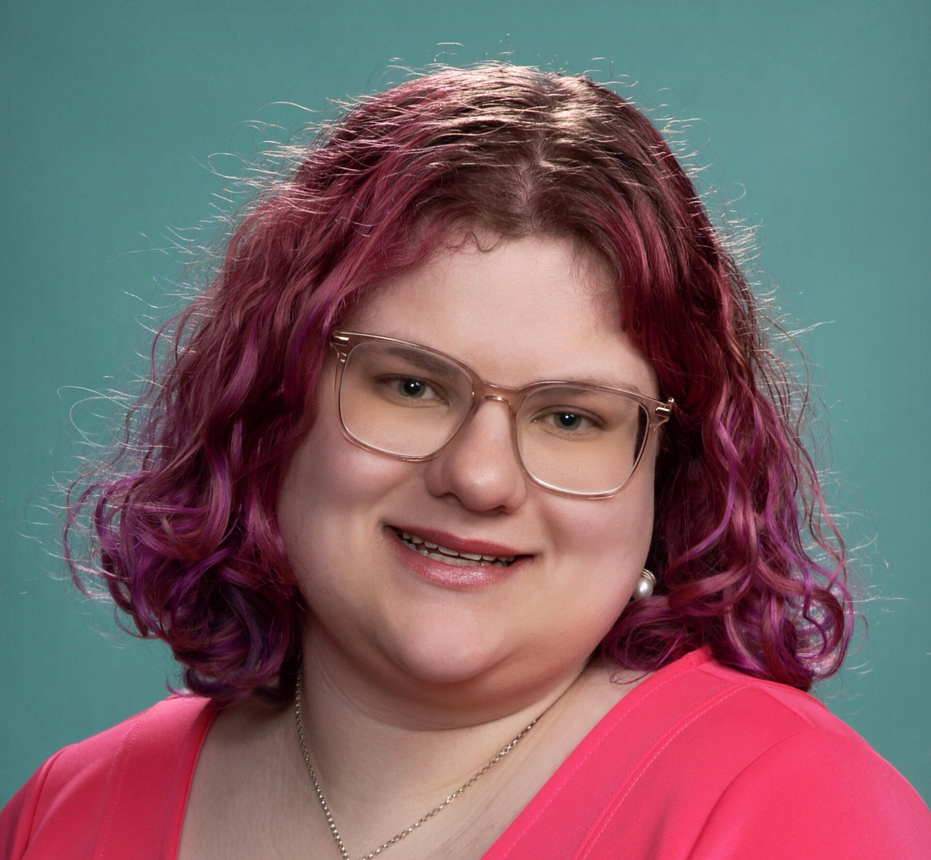 An image of Sara Slater, a white woman in her late twenties, She has blue eyes, is wearing blush pink glasses and has pink, wavy hair, She is smiling.